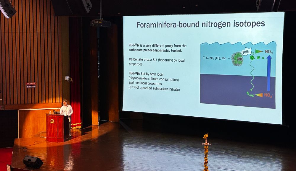 A picture of Dr. Jesse Farmer on stage at ICP15 with him in the corner left spotlight and a screen behind him with a slide that is titled "Foraminifera-bound nitrogen isotopes" 