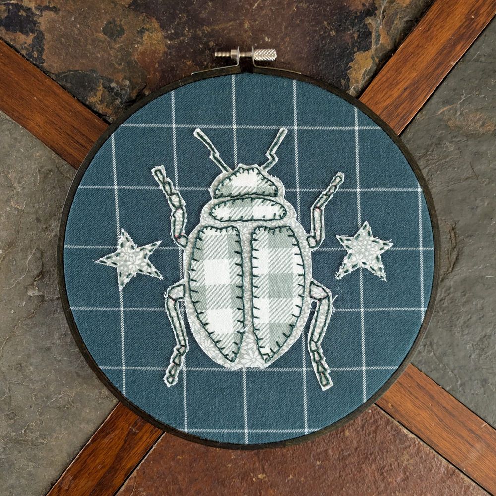A light green beetle in patterned fabric appliqued by hand onto dark green pattern fabric. The piece is framed in a dark brown embroidery hoop.