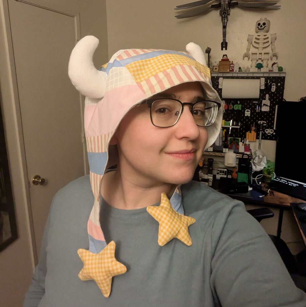 AJ wearing a hat they sewed. It is a cap made of patchwork featuring pastel pink, yellow, blue, and white strips of scrap fabric. The hat has long ear flaps with plush stars on the ends. White plush horns are seen on the head.