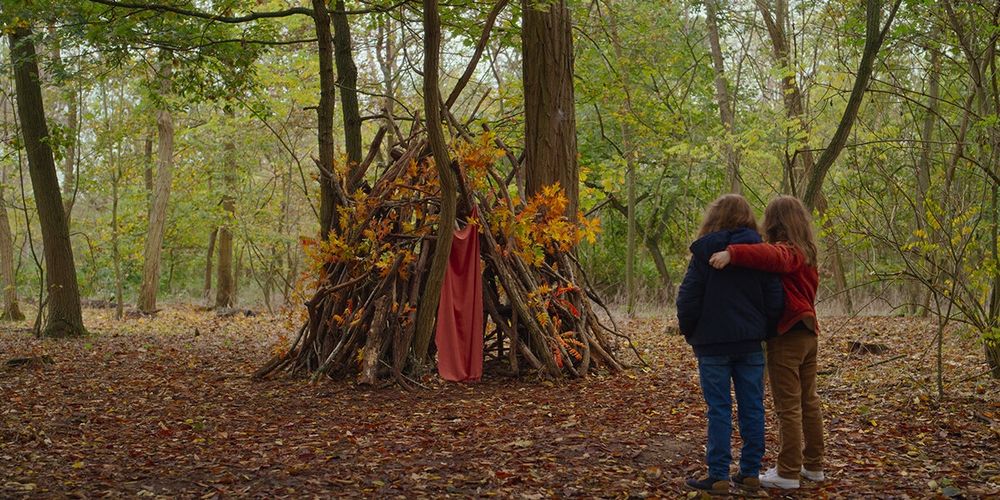A young girl and her mother (who, for reasons, are both the same age) look with pride at their fort in the woods.