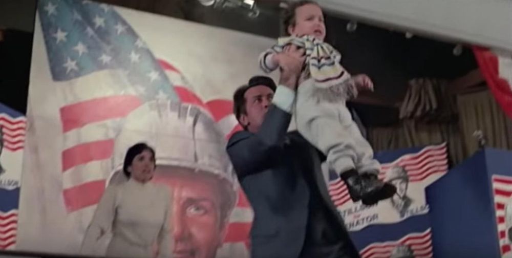 A pivotal scene from The Dead Zone shows Martin Sheen’s character, Greg Stillson, in a moment of panic as he lifts a crying baby above his head to use as a human shield against an assassin. They're on a stage decorated with American flags and campaign banners, with a giant mural of Stillson in a hardhat looming behind them. A horrified woman rushes toward him. The scene starkly contrasts political theatrics with cowardice, capturing a climactic moment of moral collapse.