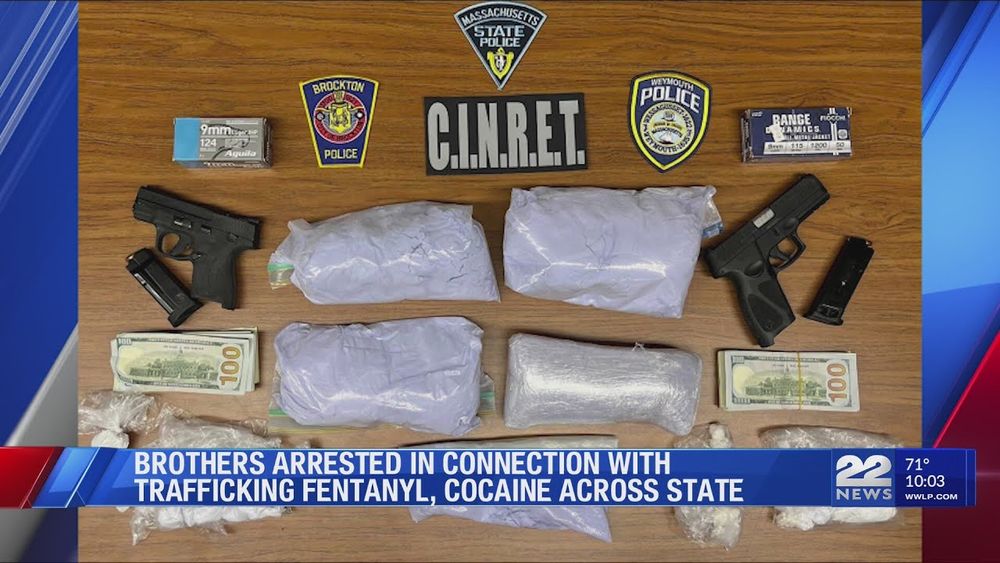 A news graphic from WWLP 22 News shows a wooden table covered with evidence from a drug trafficking arrest. Displayed are two handguns with magazines, two boxes of 9mm ammunition, several large bundles wrapped in plastic (presumably narcotics), multiple stacks of $100 bills, and law enforcement patches from Massachusetts State Police, Brockton Police, Weymouth Police, and a C.I.N.R.E.T. task force placard. The news banner reads: “BROTHERS ARRESTED IN CONNECTION WITH TRAFFICKING FENTANYL, COCAINE ACROSS STATE.”