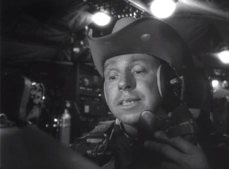 Black-and-white film still of actor Slim Pickens as Major T.J. "King" Kong in Dr. Strangelove. He's wearing a cowboy hat over a flight helmet and speaking into a radio headset, seated inside the cockpit of a B-52 bomber. The scene is dimly lit, with glowing overhead lights and detailed aircraft controls surrounding him.