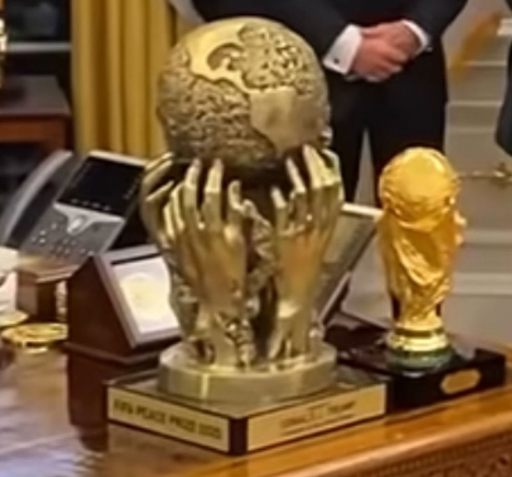 Two large gold-colored trophies sit prominently on the Resolute Desk in the Oval Office. The trophy on the left, labeled “FIFA Peace Prize 2023,” features a shiny, globe-shaped top supported by multiple sculpted hands reaching upward — an elaborate, overly ornate design that appears visually heavy and cluttered. To its right is a replica of the FIFA World Cup trophy, with a similarly glossy, exaggerated golden finish.