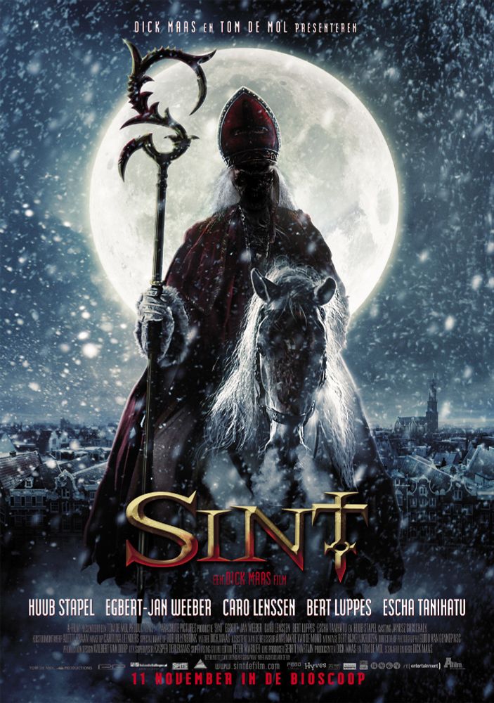 A dark, snowy movie poster for the horror film “Sint” features a menacing figure dressed like a sinister version of Saint Nicholas. He sits on a white horse, holding a jagged, spiked staff, with his face obscured in shadow beneath a tall red bishop’s hat. A giant full moon glows behind him, illuminating the rooftops of a European city below. Snow swirls through the night sky. The title “SINT” appears in large gold letters near the bottom, with cast and crew credits beneath. Text indicates that the film is directed by Dick Maas.