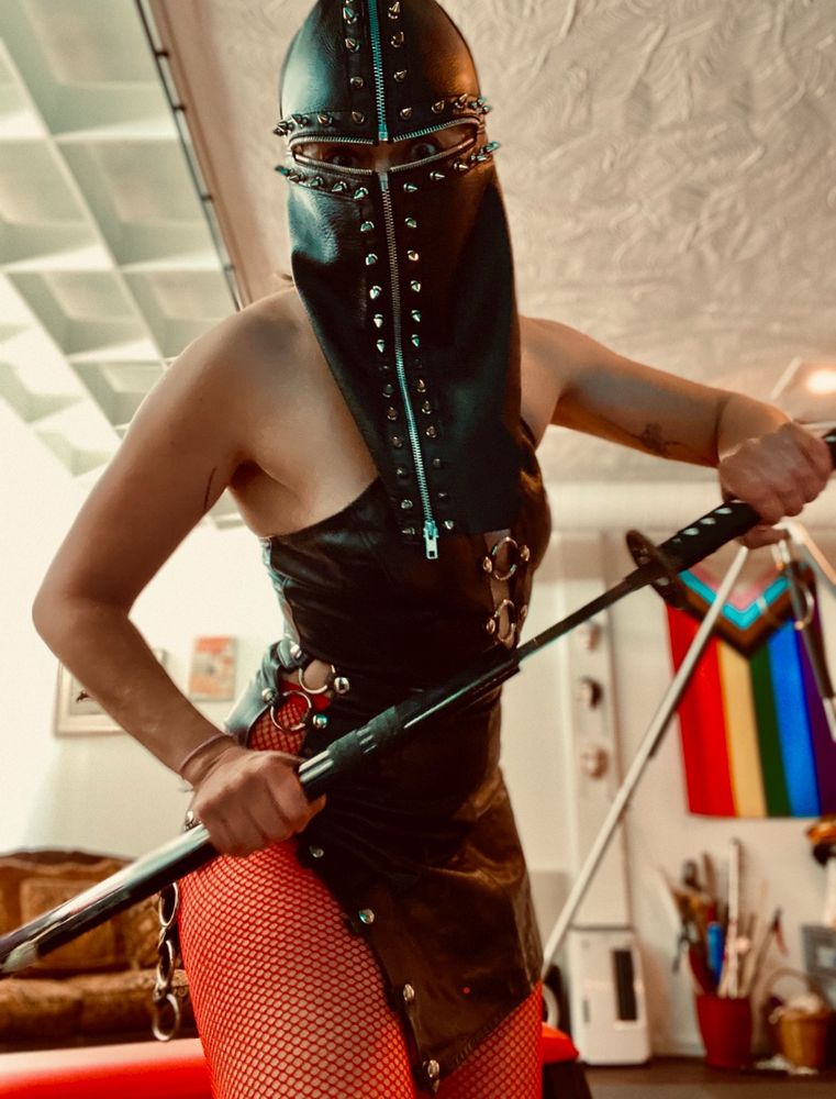 A woman, standing, with a partially-unsheathed katana, wearing a tight leather skirt and top, with a studded leather hood on