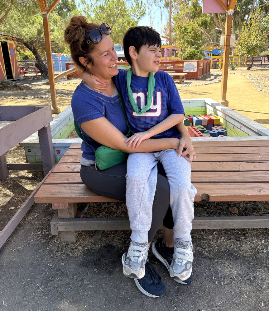 a child sits on an adults lap on a picnic bench in a park.