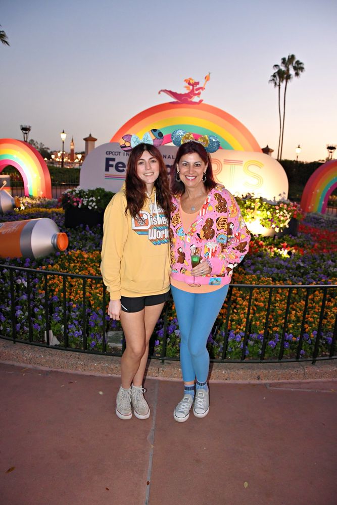 Photo shows 2 women posing in front of Epcot Center Festival of the Arts logo at sunset. 