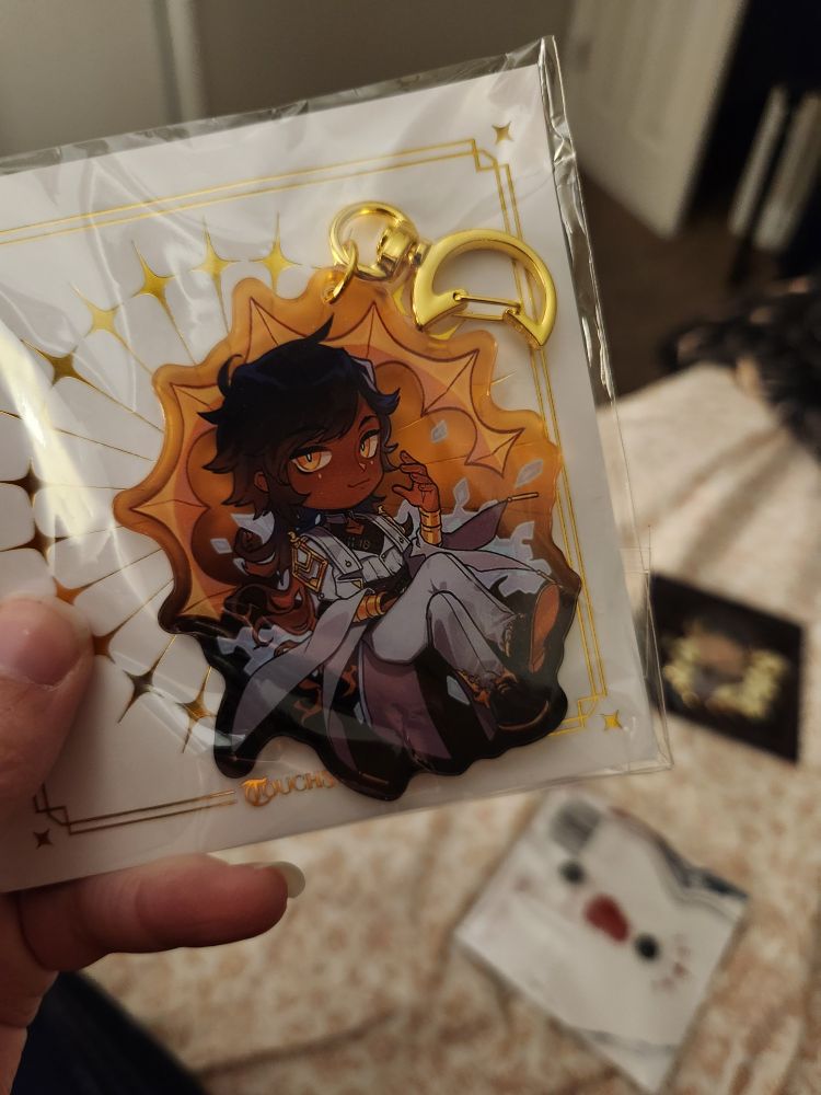A photo of my left hand holding up an acrylic charm keychain of Kuras from the game Touchstarved, inside the packaging.