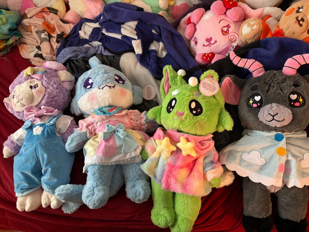Four plushies laying on a bed with a red comforter. From left to right: a purple sheep wearing a white shirt and overalls, a blue whale wearing a pink shirt and multicolored overall skirt, a green alien cat wearing a colorful fluffy jacket with a scarf with stars on the end, and a baphomet wearing a cloud cloak