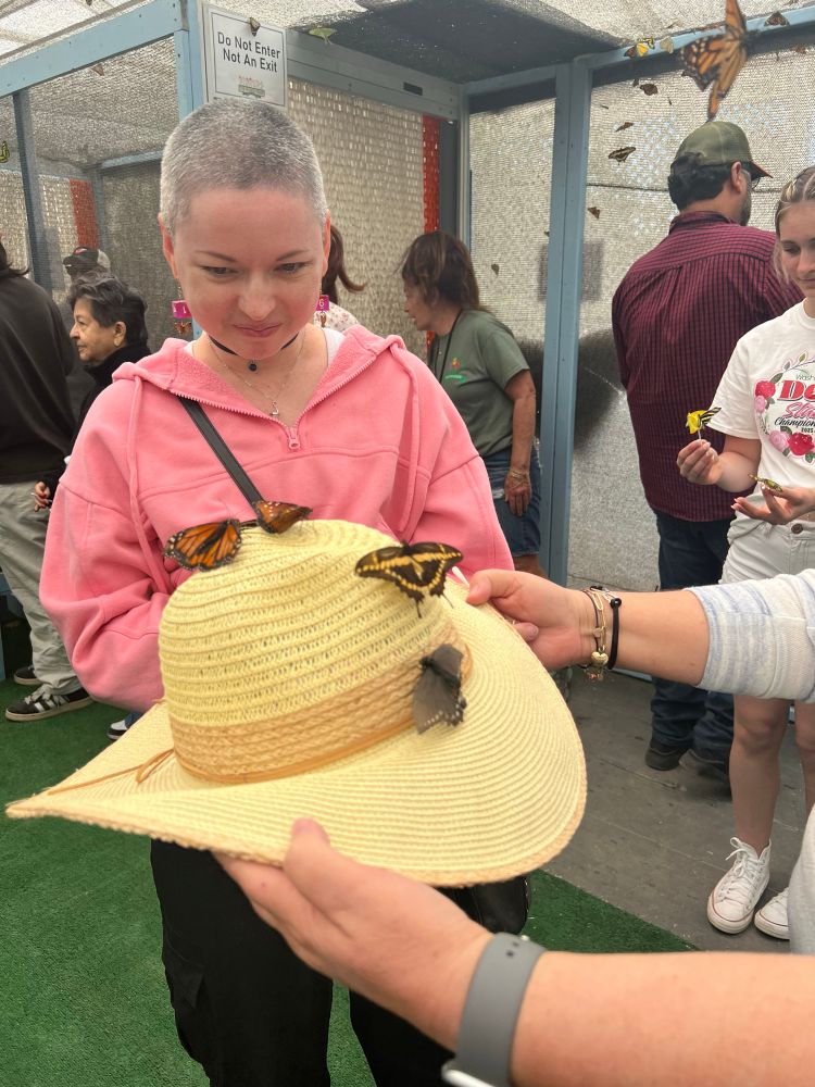 Photo of Sarah in a pink hoodie looking at her sun hat covered butterflies as someone holds out the hat in front of her.  
