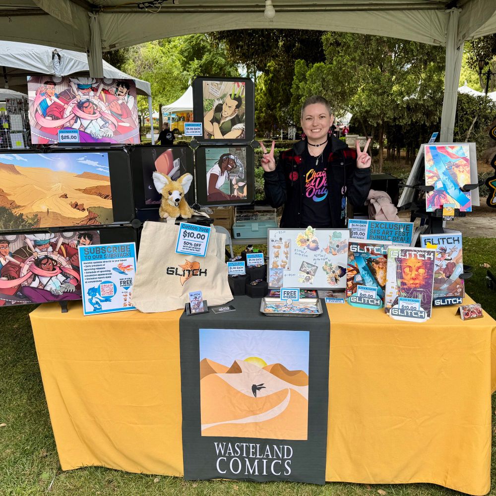 A photo of Sarah giving two peace signs while wearing a black t-shirt that reads “Chaotic Good Vibes Only” in rainbow print and a denim/plaid hoodie on top. She’s at a table with a gold tablecloth and a black table runner with the Wasteland Comics logo on it. The table is full of GLITCH comics and merchandise. 