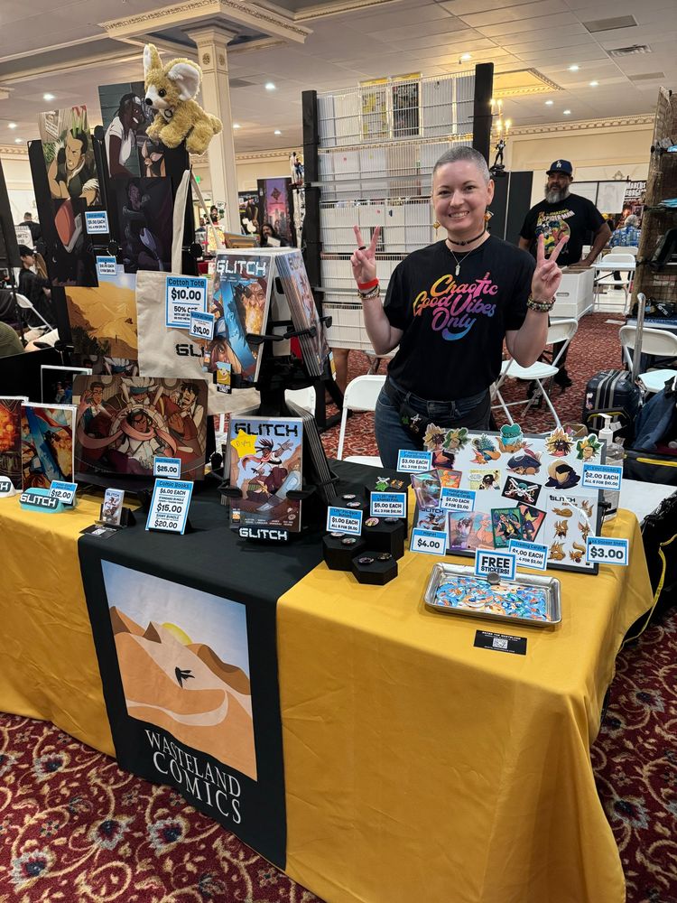 Photo of Sarah wearing a black shirt with rainbow print saying “Chaotic Good Vibes Only” while standing behind a table of Wasteland Comics merchandise and comic books. 