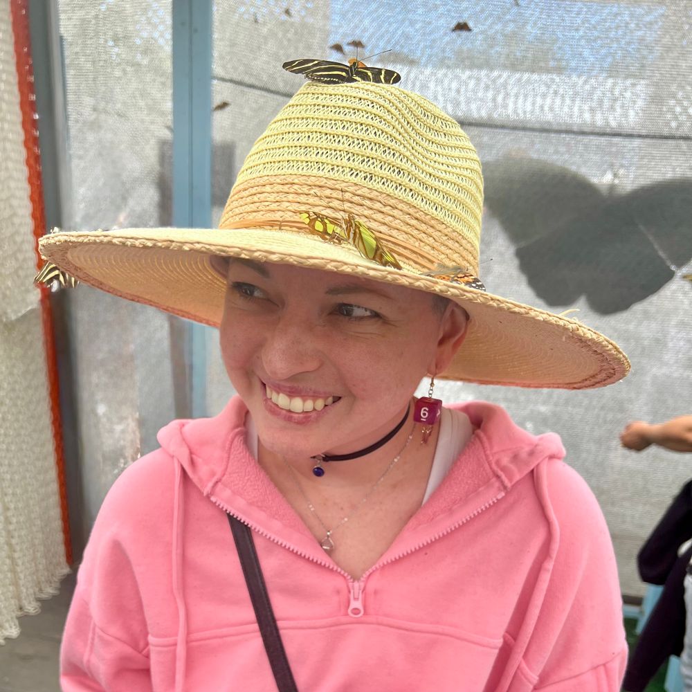 Photo of Sarah in a pink hoodie and sun hat, with butterflies resting on the sun hat. 