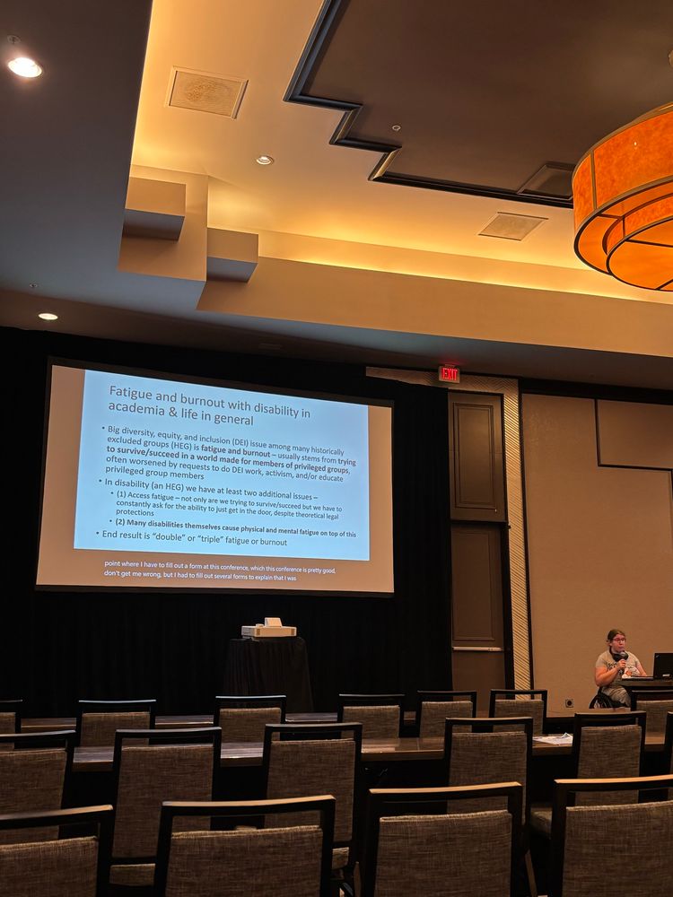 A white person in a wheelchair is speaking at a podium in front of slides. The slides heading states “Fatigue and burnout with disability in academia & life in general”