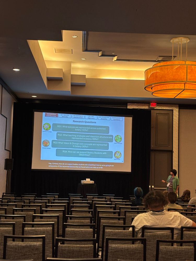 Dark short haired person stands at the front of a presentation. A slide showcases the four main questions of their talk (which I actually can’t read clearly)