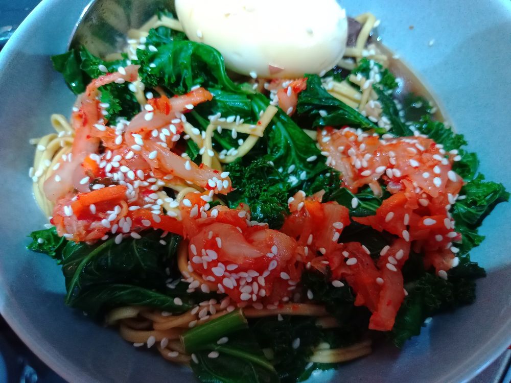 Noodles, hard boiled egg, kimchi, kale in chicken broth with sesame seeds on top.