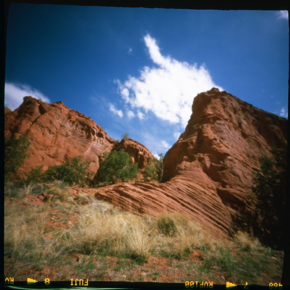 Scan of a film photograph with red Navajo Sandstone rocks showing large scale crossbedding under a blue sky with white clouds.  