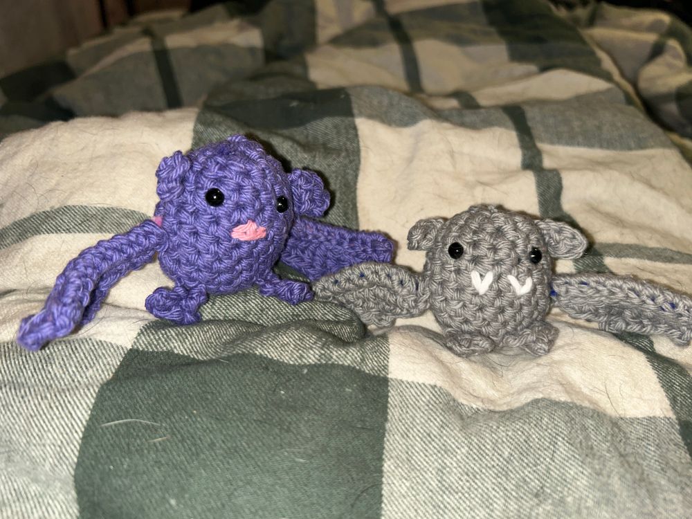 Two amigurumi bats. One is grey with white vampire fangs and the other is purple with a pink bat nose