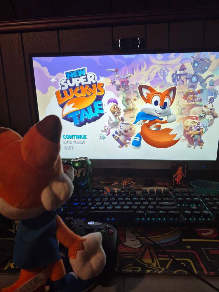 A plushie of Lucky from Super Lucky's Tale standing on a table in front of a gaming controller and computer running the game New Super Lucky's Tale.