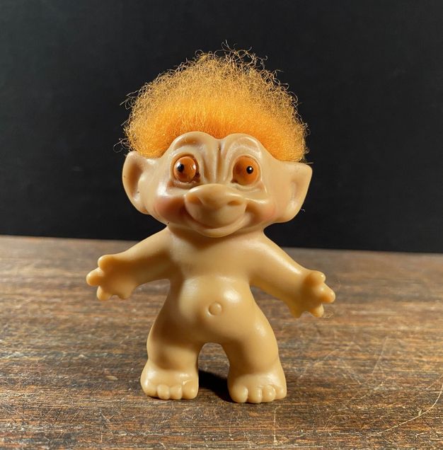 Troll doll from the 1960s - a small beige rubber/plastic doll, unclothed, with spiky furry orange hair & bulging orange eyes, looking a bit like a cheerful precursor of Yoda