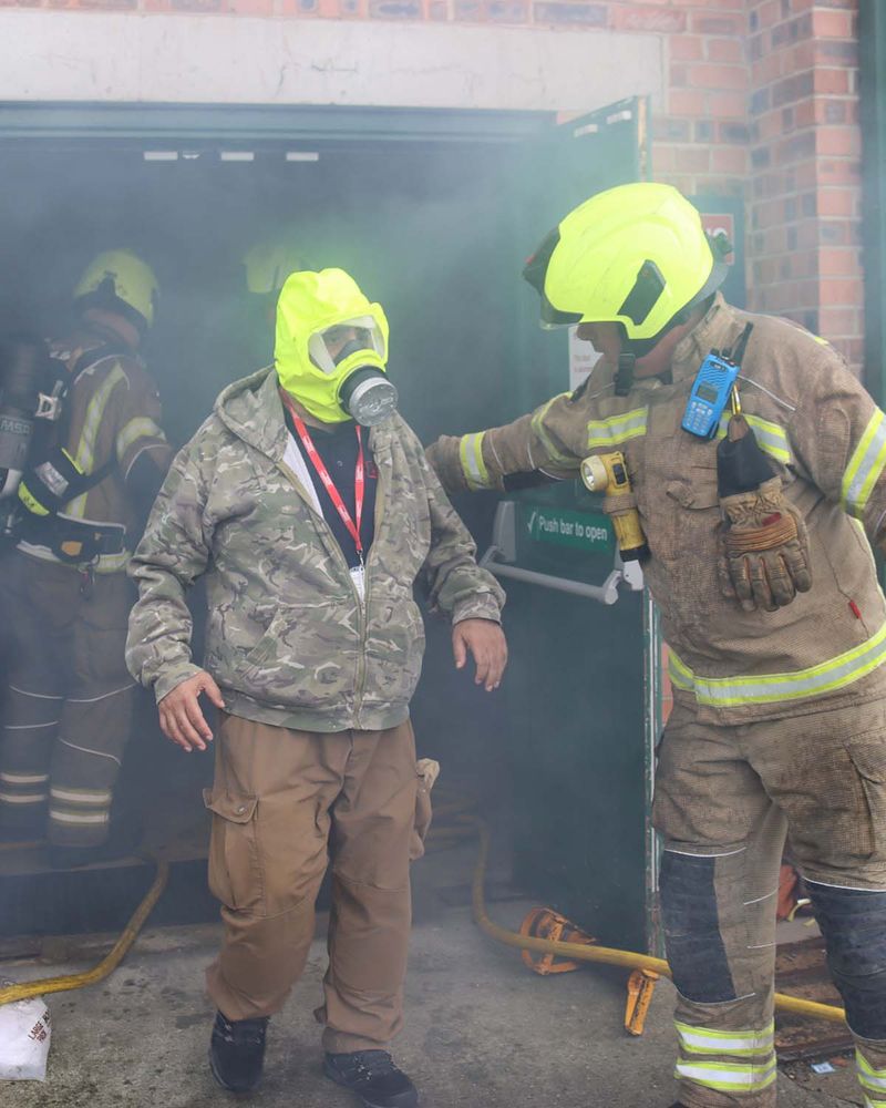 A firefighter dressed in full gear, including a neon-yellow helmet and breathing apparatus, assists a person wearing a camouflage jacket and yellow smoke hood out of a smoky building. The scene is filled with smoke, and additional firefighters are visible in the background.
