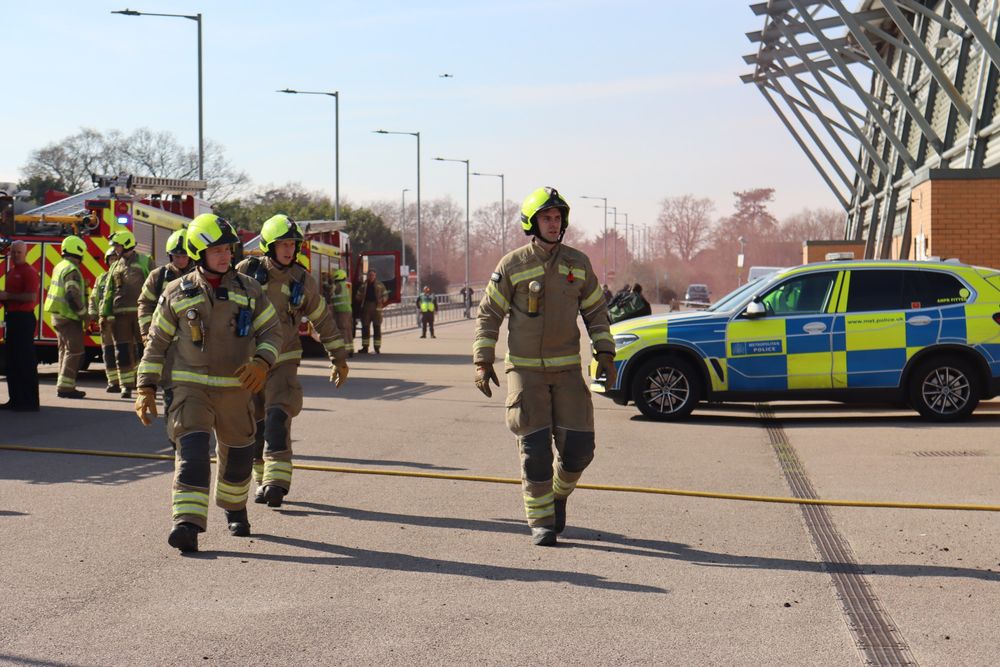 Firefighters in full gear walk past emergency vehicles, including fire engines and a marked police SUV, during a coordinated response exercise outside a large building. 