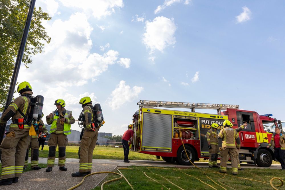 Firefighters in PPE and breathing apparatus stand together, with fire engine in background and hoses in foreground.