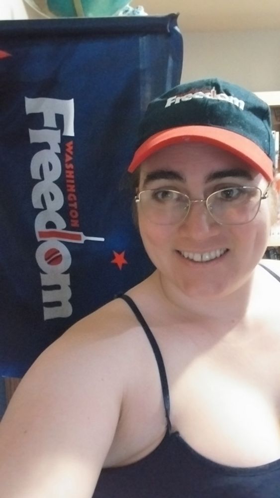 Me wearing a blue tank top and a baseball cap with the logo of the cricket team, Washington Freedom, standing in front of a Washington Freedom flag.