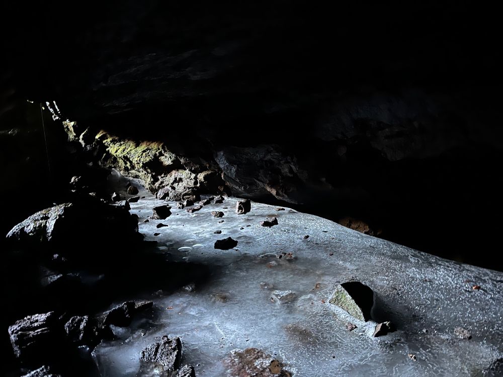 Rocks jut out from a sheet of cloudy ice at the bottom of a cave, lit up by sunlight from the entrance at top left. Some green moss is visible on the wall in the background. 