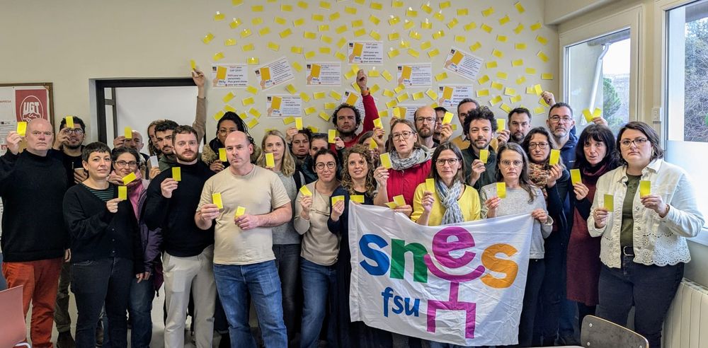 photo de groupe des personnels du LFB, en salle des professeur·es, brandissant un carton jaune pour dénoncer l'absence de dialogue social à l'AEFE. Un drapeau SNES-FSU au premier plan