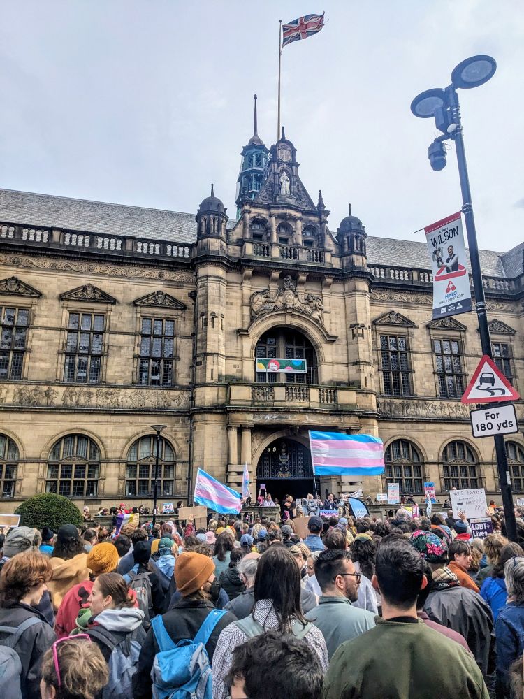 A photo of thousands of people gathered outside an old fashioned building at a protest, with some holding trans pride flags