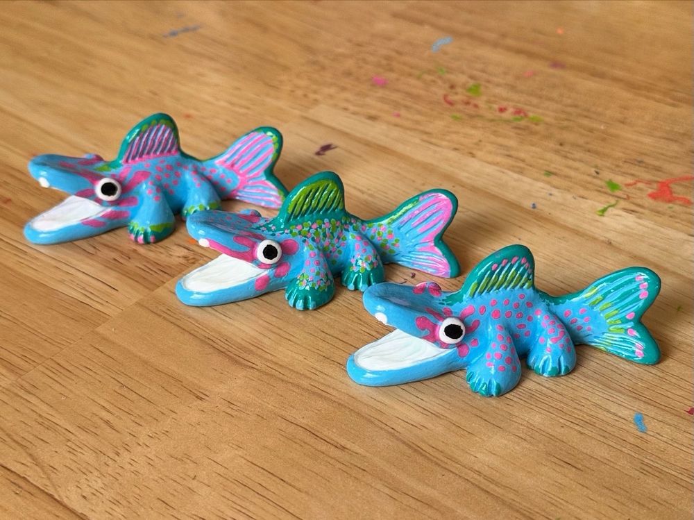 Photo of three fish creature figurines on a table. Their bodies are painted with various patterns using blue, green, and pink