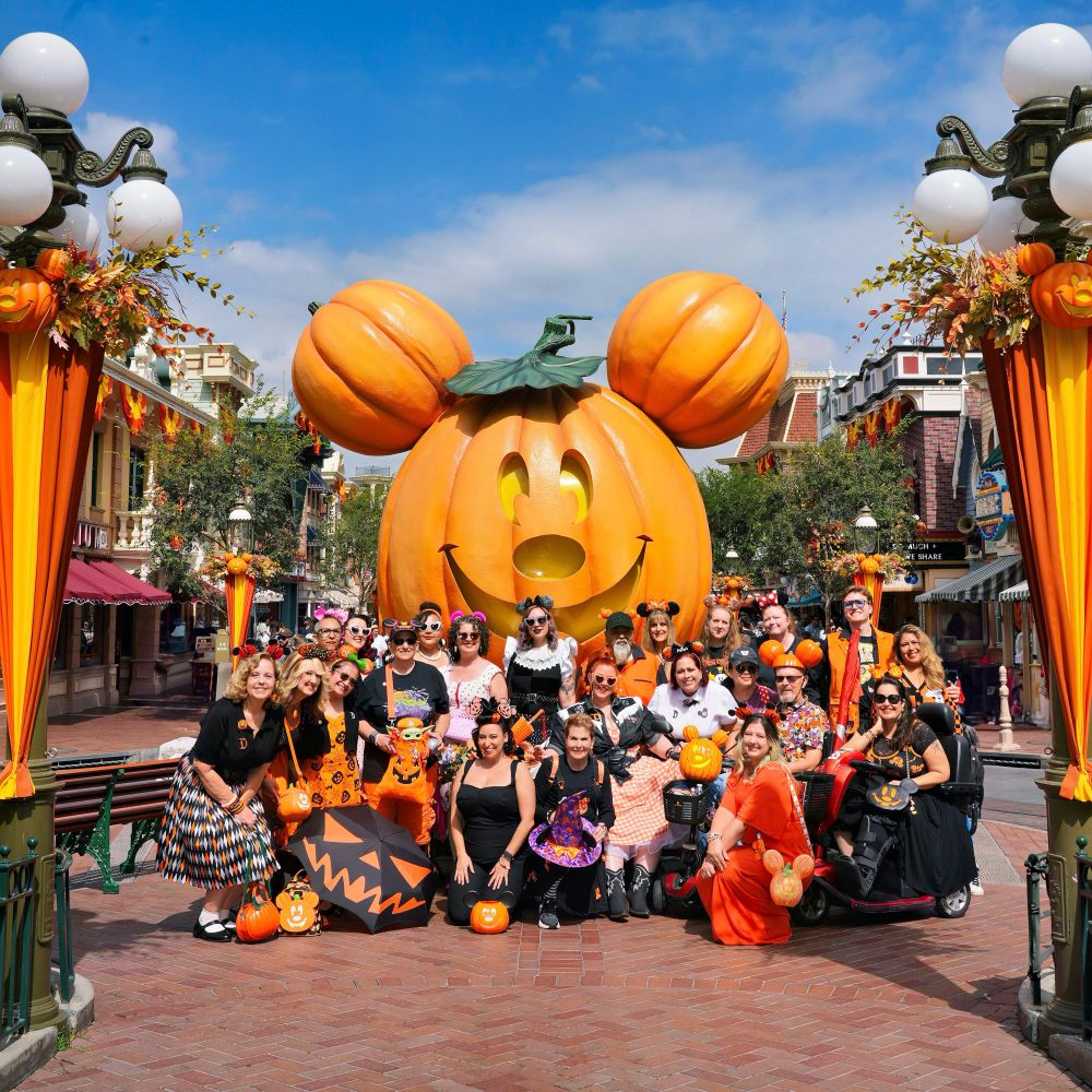 A group of people dressed in festive Halloween outfits pose together in front of the giant Mickey pumpkin on Main Street at Disneyland. The group wears a mix of orange, black, and seasonal clothing with pumpkin patterns, mouse ears, parasols, and themed accessories. Some hold pumpkin buckets or umbrellas with jack-o’-lantern faces, while a few ride decorated mobility scooters. The background shows Main Street buildings and lamp posts draped in orange and yellow fall garlands.