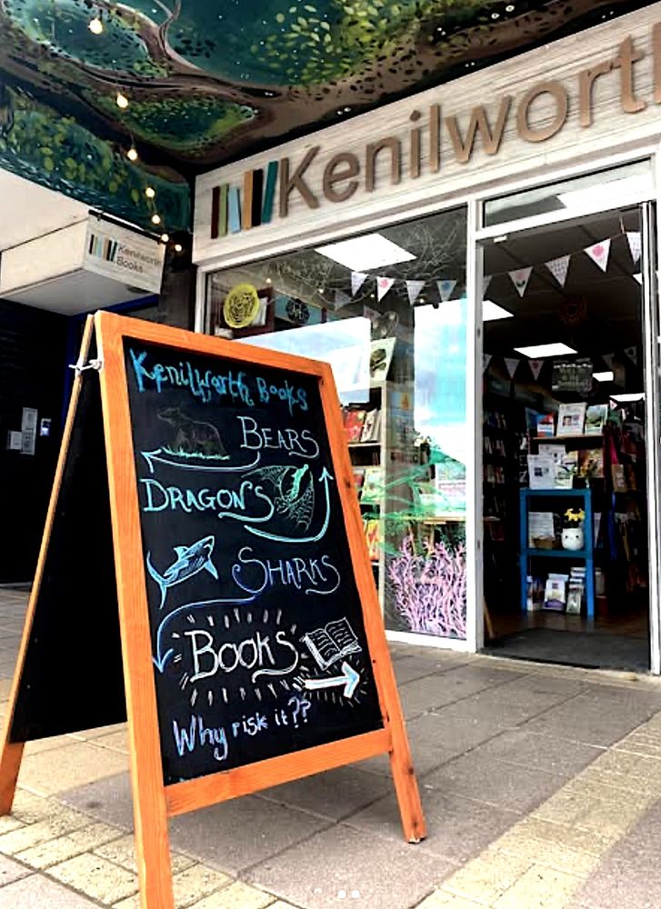 The front of the bookshop (which has bunting hanging inside) with a chalkboard sign outside saying: Bears, Dragons, Sharks, Books, why risk it?
