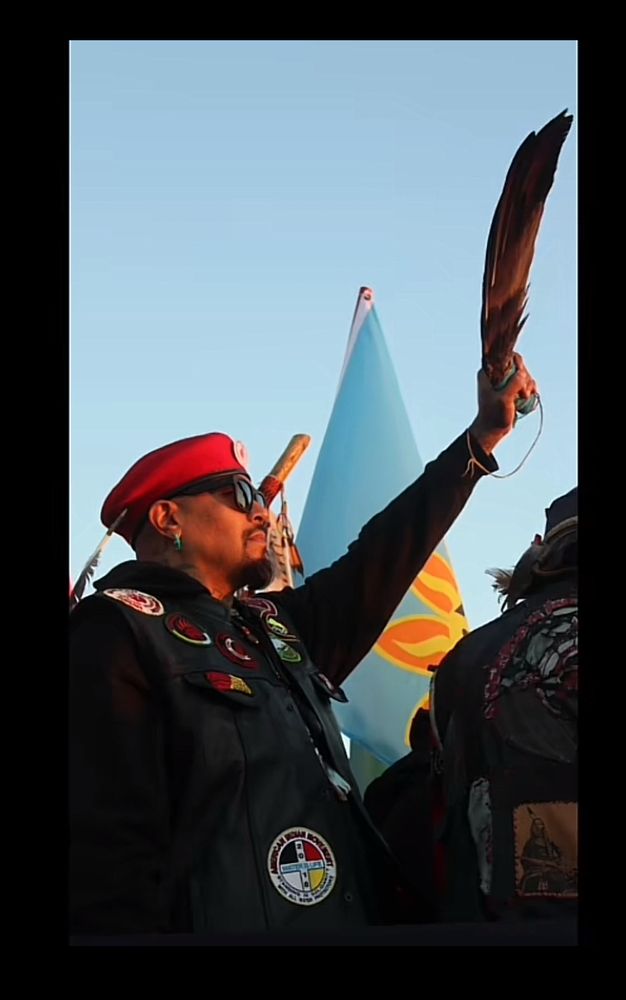 sam, a native man with brown skin and a tall build, wearing an aim vest and red hat, and sunglasses holding up an eagle feather wing with the blue sky behind him