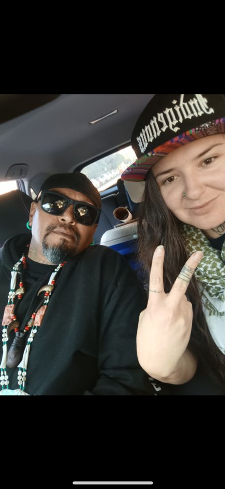 sam with a baseball cap, medicine (necklaces), sunglasses in the passenger seat sitting next to me throwing up a peace sign wresting a hat that says indigenous. a cooler and poster are visible in the background back seat 