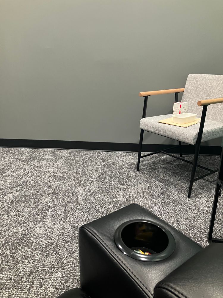 An uncomfy-looking grey and black chair in the corner of a room with grey carpet and grey walls with black trim. A manilla folder and a white sprovato box are on the chair