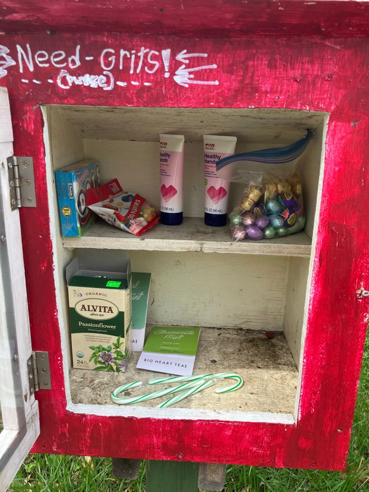 A grimy little free library stocked with snacks. Dots, Jelly Bellys, two tubes of hand lotion, a ziplock bag of lil colorful chocolate eggs and bunnies, some boxes of tea, and three green and white candy canes