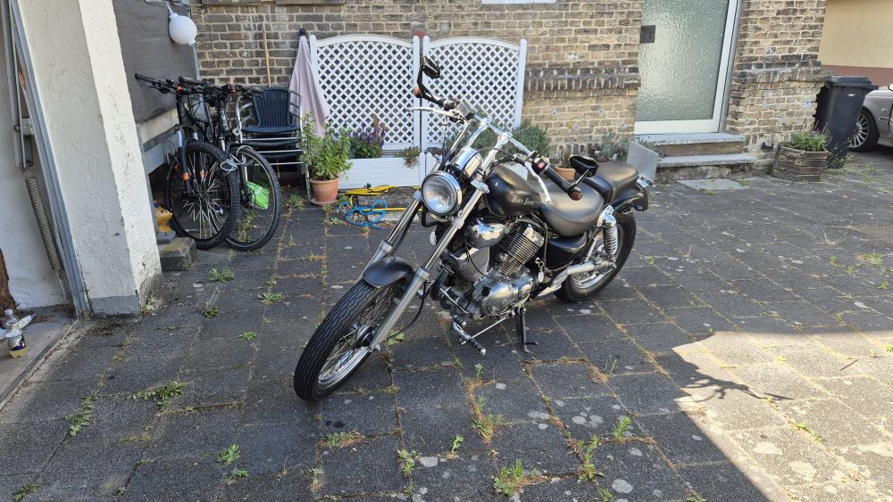 Schwarzes Custom Bike mit V-Motor und Chromteilen, basierend auf einer Yamaha Virago, schräg geparkt auf einem gepflasterten Hof vor einer Backsteinwand mit Pflanzen und Fahrrädern im Hintergrund.