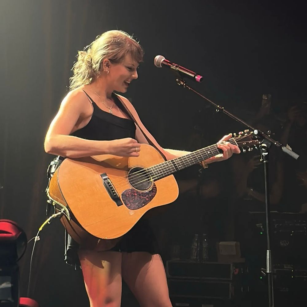 Taylor Swift performing “Shake it off” at Tight Ends with an acoustic guitar. She is wearing a plain black dress.