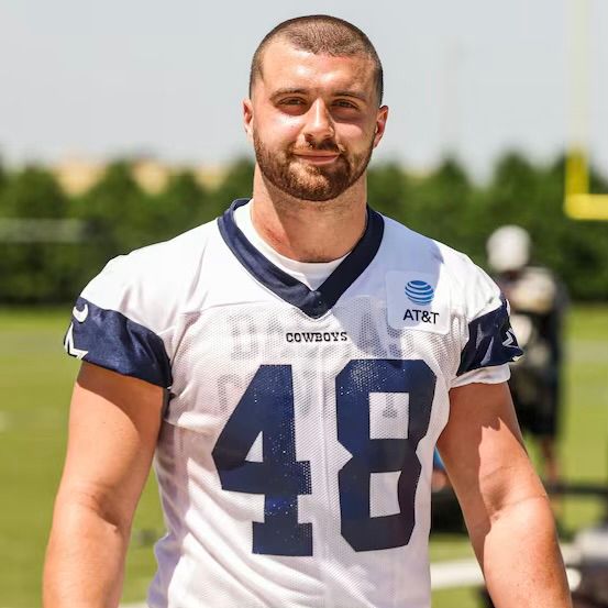 Dallas Cowboys Jake Ferguson in his white Jersey with the number 48 printed on Dakr blue. Background is a grassy field and blurred.