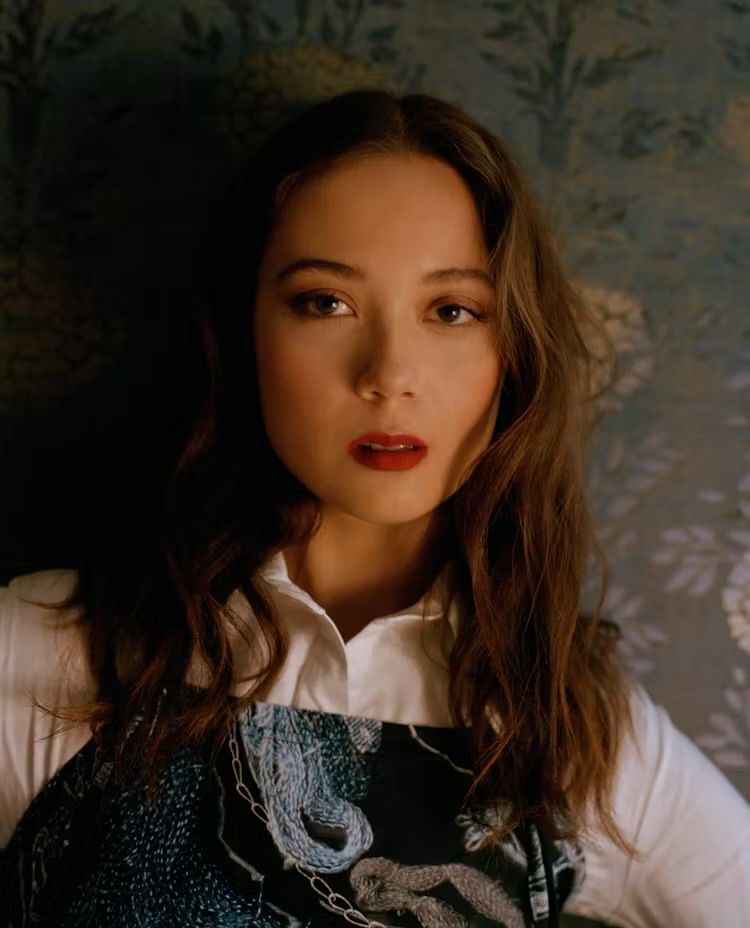 Laufey wearing a white shirt and black overalls with a blue a grey prinr. She is standing infront of a wall with free wallpaper with a flower pattern.
