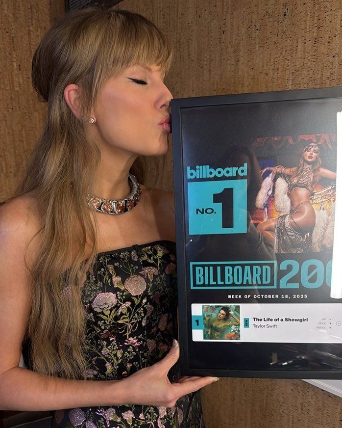 Taylor Swift wearing a black dress with floral patterns and a thin silver and bronze chocker. She is kissing a frame award for her album “life of a Showgirl,” being number one on Bilboard.