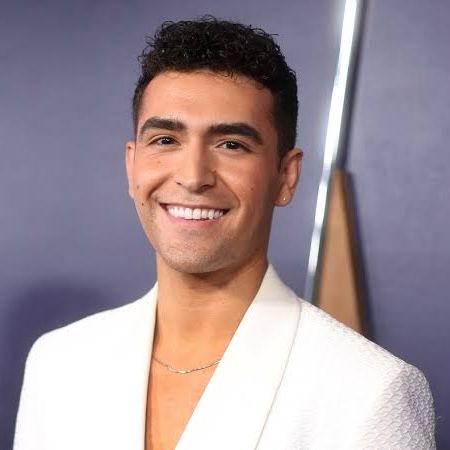 Photo of Ezra Sosa wearing a white suit jacket and silver necklace. He is smiling. The background is a light purple wall