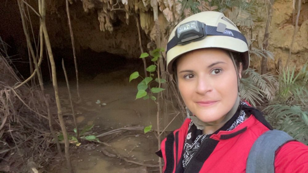 Eu, de capacete com lanterna e macacão vermelho em frente a uma caverna.