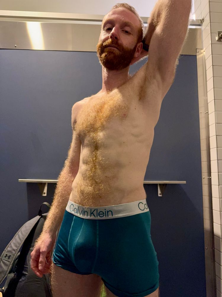 Hair toned ginger man standing in change room wearing blue Calvin Klein trunks, left arm raised above head