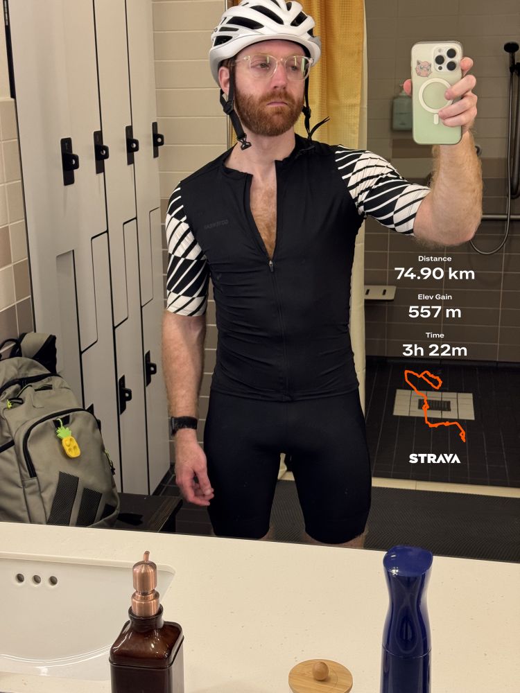 Bearded ginger man standing in locker room mirror in tight cycling gear. Strava stats listed (74.9km, 557 elevation gain, 3 hours 22 minutes)