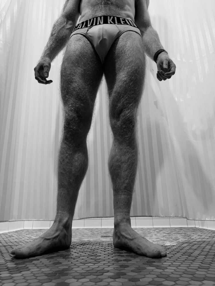 Black and white photograph of a hairy man standing in front of a shower curtain. Feet to waist are in frame, with light colored Calvin Klein briefs with a mild print showing through. 