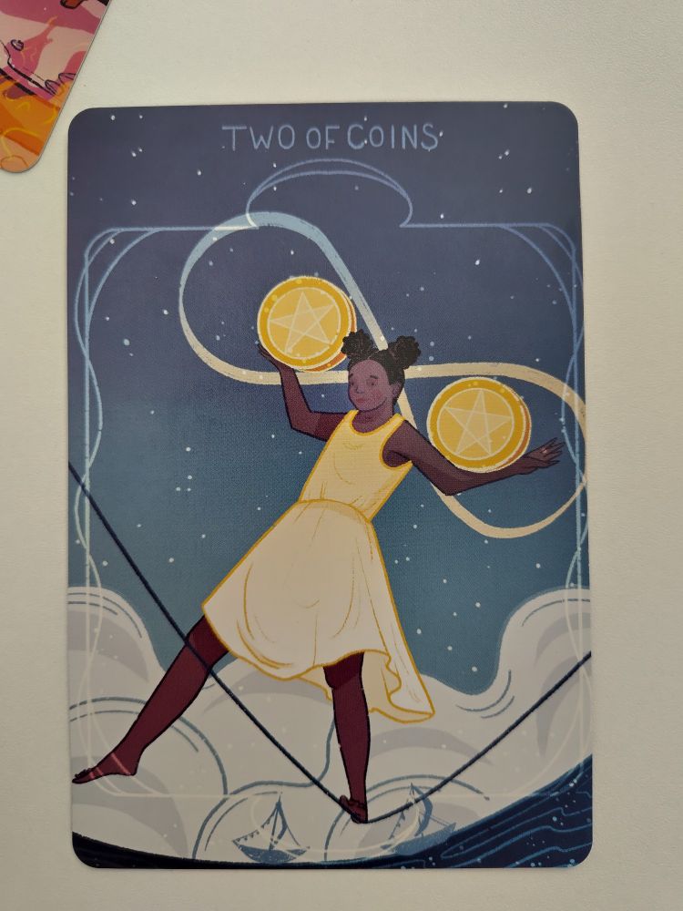 Single tarot card of the two of coins with central figure in yellow dress balancing on one foot on a tightrope, holding yellow circles with five pointed stars on the front, one in the crook of her elbow, the other on her other hand